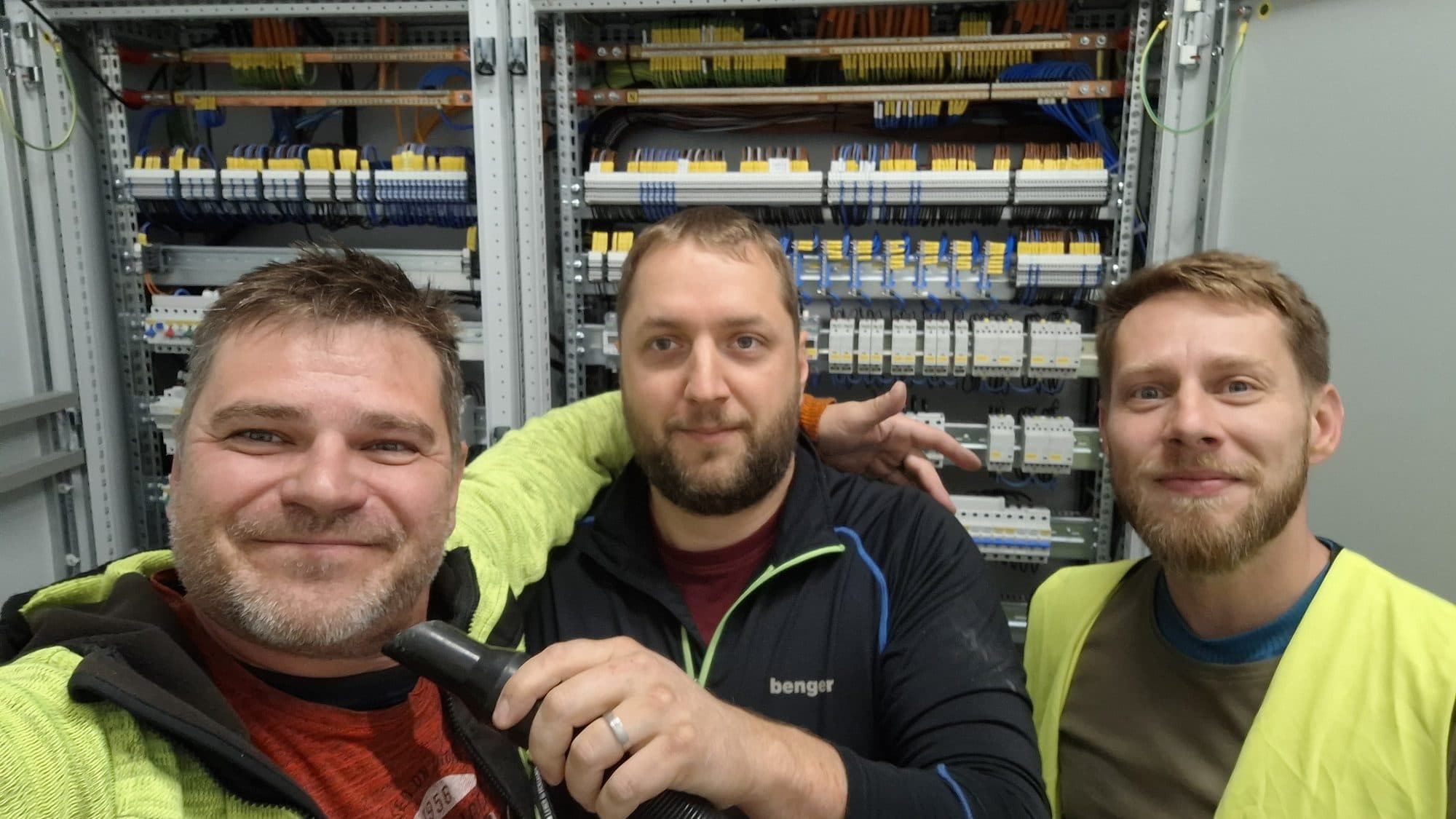 Tři členové týmu OKelectric se usmívají před velkým rozvaděčem